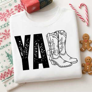 Puede incluir: Una sudadera blanca de cuello redondo con el texto negro Y'ALL, donde las letras L están sustituidas por un par de botas vaqueras dibujadas con líneas. Las botas están decoradas con un pequeño lazo en el lateral. El diseño está centrado.