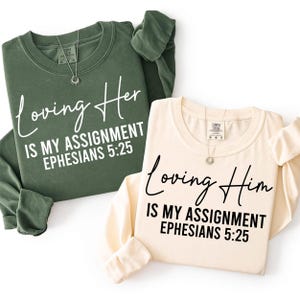 May include: Two long-sleeved sweatshirts, one olive green and one cream, with the text "Loving Her is My Assignment Ephesians 5:25" and "Loving Him is My Assignment Ephesians 5:25" in white and black script. Each shirt has a necklace.