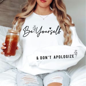 Puede incluir: Sudadera blanca con el texto "Be Yourself & DON'T APOLOGIZE" en escritura negra. La sudadera tiene un diseño floral en la parte delantera y en la manga. La persona sostiene un vaso de té helado.