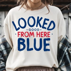 Könnte beinhalten: Cremefarbenes Sweatshirt mit dem Text "LOOKED GOOD FROM HERE BLUE" in blauen und roten Buchstaben. Das Sweatshirt hat einen Rundhalsausschnitt und lange Ärmel. Ein kariertes Hemd ist über den Schultern drapiert.