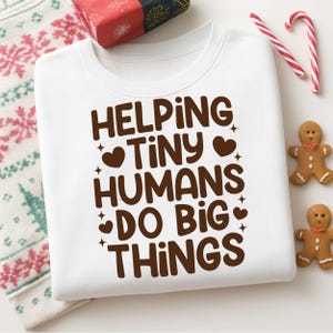 May include: White sweatshirt with the text "HELPING TINY HUMANS DO BIG THINGS" in brown. The sweatshirt is folded with a Christmas theme, including gingerbread men, candy canes, and a wrapped gift.