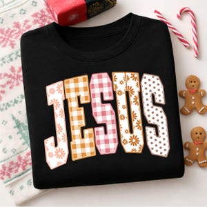 May include: A black crewneck sweatshirt featuring the word JESUS in large, block-style letters. Each letter has a unique pattern: floral, orange gingham, pink gingham, floral, and brown polka dots, arranged in a slight arch across the chest.