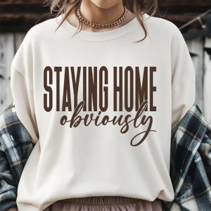 May include: A cream-coloured crewneck sweatshirt featuring the dark brown text "STAYING HOME" in a bold, sans-serif font, followed by the word "obviously" in a brown, cursive script. The garment is worn with a dark blue and green plaid shirt.