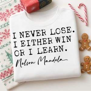 Op de afbeelding: Witte sweatshirt met de quote "I NEVER LOSE. I EITHER WIN OR I LEARN." van Nelson Mandela in zwarte tekst. De afbeelding bevat feestelijke decoraties zoals peperkoekmannetjes en zuurstokken.