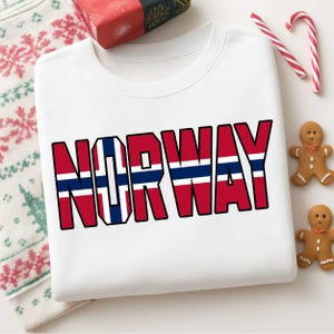 Op de afbeelding: Een wit sweatshirt met ronde hals en het woord NORWAY in grote blokletters. De letters zijn gevuld met het rood, wit en blauwe patroon van de Noorse vlag. Het sweatshirt ligt op een plat oppervlak met kerstdecoratie.