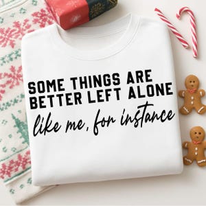 Puede incluir: Sudadera blanca con el texto negro "SOME THINGS ARE BETTER LEFT ALONE like me, for instance". La imagen incluye un regalo envuelto, bastones de caramelo y hombres de jengibre, lo que sugiere un tema festivo.
