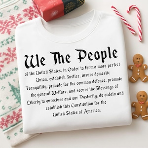 Może przedstawiać: Biała bluza z napisem "We The People" gotycką czcionką, a następnie preambułą do Konstytucji Stanów Zjednoczonych. Obraz zawiera piernikowych ludzików, laski cukrowe i zapakowany prezent, sugerując świąteczny motyw.