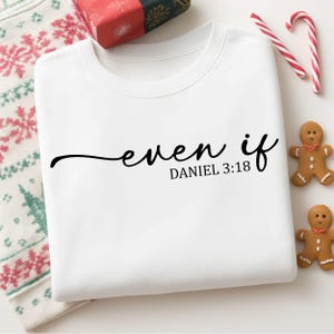 以下が含まれることがあります： 「even if」と「DANIEL 3:18」の文字が黒い筆記体で書かれた白いスウェットシャツ。 折りたたまれたスウェットシャツは、ジンジャーブレッドマン、キャンディケーン、包装されたギフトなど、お祝いの装飾に囲まれています。