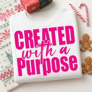 May include: A white sweatshirt with the text "CREATED with a PURPOSE" in bright pink. The image includes a wrapped gift, candy canes, and gingerbread men, suggesting a holiday theme.