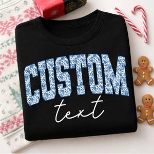 Peut inclure: Sweat-shirt noir avec le mot "CUSTOM" en grandes lettres bleues à motifs floraux. En dessous, le mot "text" est écrit en cursive. Le sweat-shirt est entouré de décorations de Noël, dont des bonhommes en pain d'épices et des cannes en sucre.