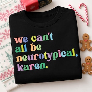 Op de afbeelding: Zwarte crewneck sweatshirt met de tekst "we can't all be neurotypical, karen" in regenboogkleuren. De sweatshirt is omgeven door kerstversieringen, waaronder peperkoekenmannetjes en zuurstokken.