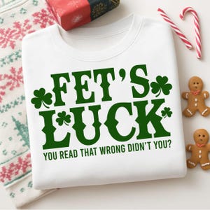 May include: White sweatshirt with the text "FET'S LUCK" in green, with shamrock accents. The phrase "YOU READ THAT WRONG DIDN'T YOU?" is below. Candy canes and gingerbread men are in the background.