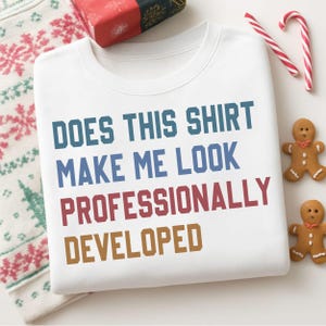 May include: A white crewneck sweatshirt featuring the text "DOES THIS SHIRT MAKE ME LOOK PROFESSIONALLY DEVELOPED" printed in blue, red, and brown block lettering. The shirt is displayed alongside gingerbread cookies and candy canes.