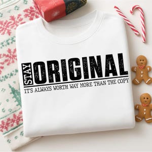 May include: White sweatshirt with the text "STAY ORIGINAL" in black block letters. The phrase "IT'S ALWAYS WORTH WAY MORE THAN THE COPY" is below. The sweatshirt is surrounded by holiday decorations, including candy canes and gingerbread men.