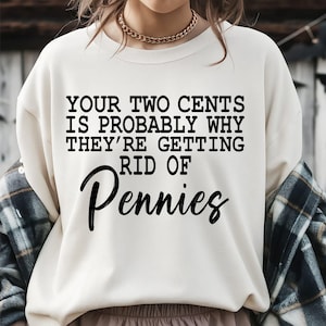 Może przedstawiać: Kremowa bluza z okrągłym dekoltem i czarnym napisem: YOUR TWO CENTS IS PROBABLY WHY THEY'RE GETTING RID OF Pennies. Odzież posiada długie rękawy i prążkowany dekolt, noszona ze złotym naszyjnikiem z łańcuszka oraz koszulą w kratę.