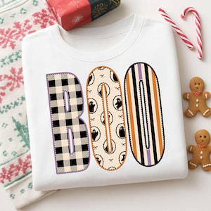 May include: A white crewneck sweatshirt featuring the word BOO printed in large letters. The B has a black and white buffalo check pattern, the O features a cream background with small ghost illustrations, and the final O has vertical stripes.