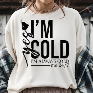 May include: Cream-colored sweatshirt with the text "I'M COLD" in large black letters. Below the text is "I'M ALWAYS COLD me 24/7". A black heart and the word "yes" are on the left side.