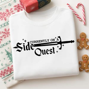Puede incluir: Una sudadera blanca de cuello redondo con un estampado gráfico negro que dice "Currently on a Side Quest" con una ilustración de una espada integrada en el texto, acentuada por pequeñas estrellas centradas en la parte delantera.