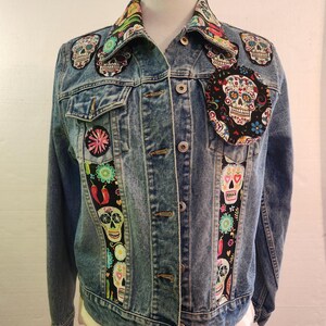 May include: A light blue denim jacket adorned with colorful skull patches and fabric accents. The jacket features a classic button-up design with a collared neckline. Patches include floral and skull designs, adding a unique, artistic flair to the garment.