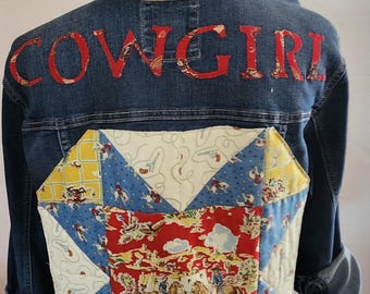 May include: Blue denim jacket with "COWGIRL" in red fabric letters across the upper back. The lower back features a patchwork design with cowboy and western-themed fabric in red, blue, and yellow.