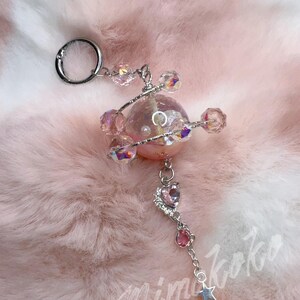May include: A pink and silver keychain with a celestial theme. The keychain features a central pink orb, surrounded by iridescent crystals and smaller pink gems. A silver chain leads to a star charm. The text "Mimikoko" is visible in the bottom right corner.