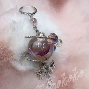 May include: A silver keychain with a clear ring, adorned with a cluster of faceted crystals and a pearlescent orb. A decorative silver butterfly charm and a teardrop crystal dangle below the orb. The keychain rests on a fluffy, pink surface.
