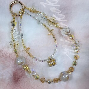 May include: A gold-toned keychain with three beaded strands. The strands feature clear, gold, and iridescent beads, with decorative elements like butterflies and a four-leaf clover. The background is a soft, pink and white surface.