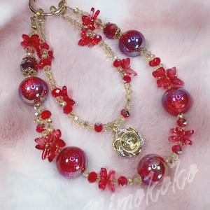 May include: A beaded lanyard featuring red butterfly-shaped beads, iridescent red spherical beads, and smaller translucent gold beads. The accessory includes a gold-toned metal ring at the top and a circular gold-toned charm with a central crystal.