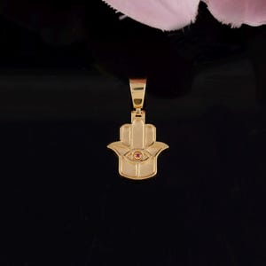 May include: Gold-colored Hamsa hand pendant with a small red gemstone in the center of the eye. The pendant is suspended from a matching gold bail. The background is black.