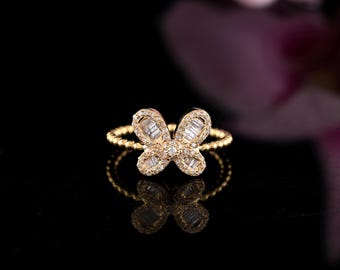 May include: A gold butterfly ring with a textured band. The butterfly design is encrusted with small, sparkling stones. The ring is set against a black background with a blurred pink flower in the background.