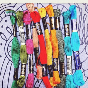 May include: A collection of colorful embroidery floss in shades of green, pink, orange, yellow, and blue. The floss is arranged on a patterned gray background, perfect for various needlework projects.