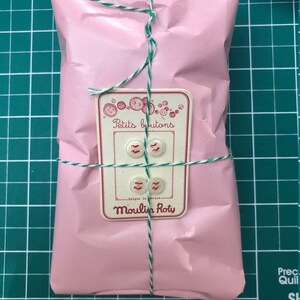 May include: A pink package wrapped in tissue paper and tied with white and green twine. A white label with red lettering reads "Petits boutons - Exigez la marque - Moulin Roty".