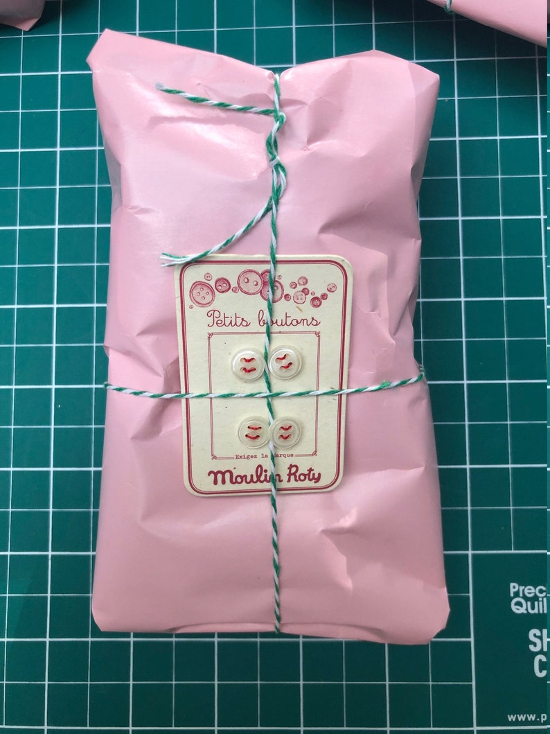 May include: A pink package wrapped in tissue paper and tied with white and green twine. A white label with red lettering reads "Petits boutons - Exigez la marque - Moulin Roty".