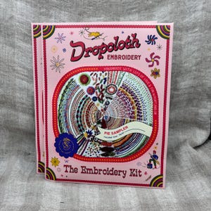 May include: A pink embroidery kit titled "Dropcloth Embroidery" with a "Pie Sampler" design. The kit features colorful embroidery patterns and the text "Handmade with lots of love & fun in Portland, OR". The finished size is 16x16 inches.