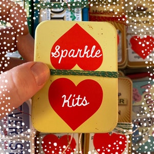May include: A yellow tin with a red heart design. The tin says "Sparkle Kits" with the word "Sparkle" in white and the word "Kits" in white inside the heart.