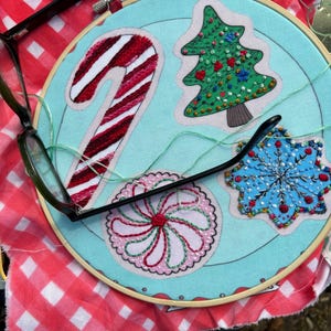 May include: A blue embroidery hoop with a candy cane, a Christmas tree, a snowflake, and a peppermint swirl design. The hoop is on a red and white checkered fabric.