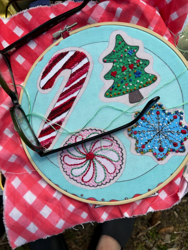 May include: A blue embroidery hoop with a candy cane, a Christmas tree, a snowflake, and a peppermint swirl design. The hoop is on a red and white checkered fabric.