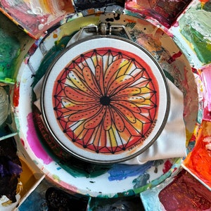 May include: A round embroidery hoop with a colorful, geometric design in shades of orange, yellow, and black. The design resembles a stylized flower or sunburst. The text "PREFECTH SAMPLERS" is printed on the fabric.