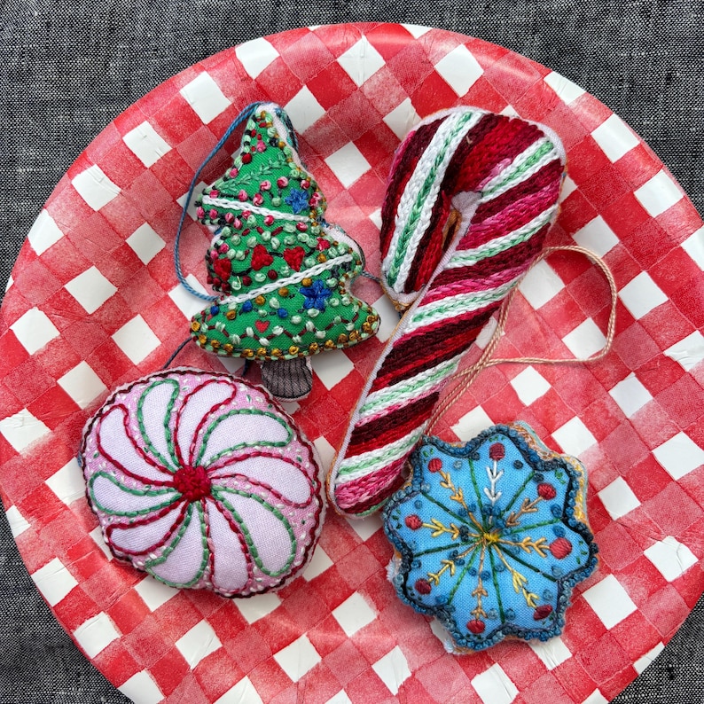 May include: Four handmade Christmas ornaments on a red and white checkered background. The ornaments are a green Christmas tree, a candy cane, a pink and white peppermint swirl, and a blue snowflake.