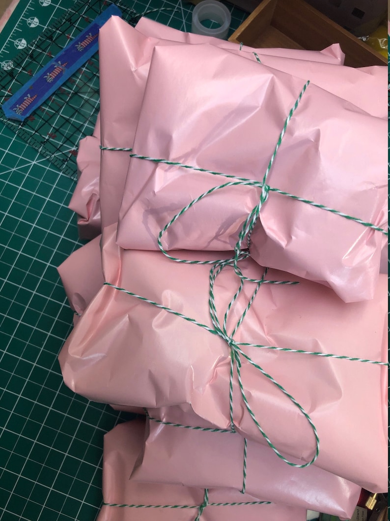 May include: A stack of pink gift bags tied with white and green twine. The bags are wrapped in a shiny, crinkled paper.
