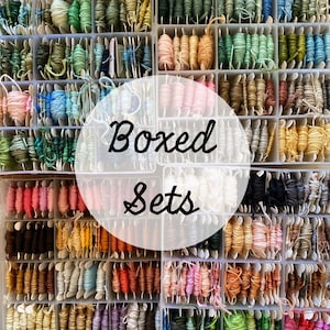 May include: A grid of small plastic boxes filled with colorful embroidery floss. The floss is in various shades of green, blue, pink, orange, and brown. The text "Boxed Sets" is overlaid on the image in a white circle.