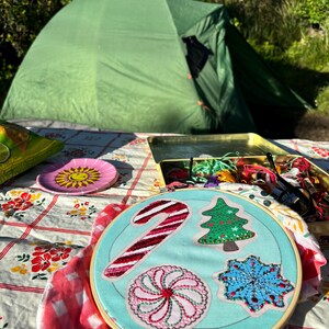 May include: A close-up of a blue embroidery hoop with a candy cane, a Christmas tree, a peppermint swirl, and a snowflake design. The hoop is on a red and white checkered tablecloth.
