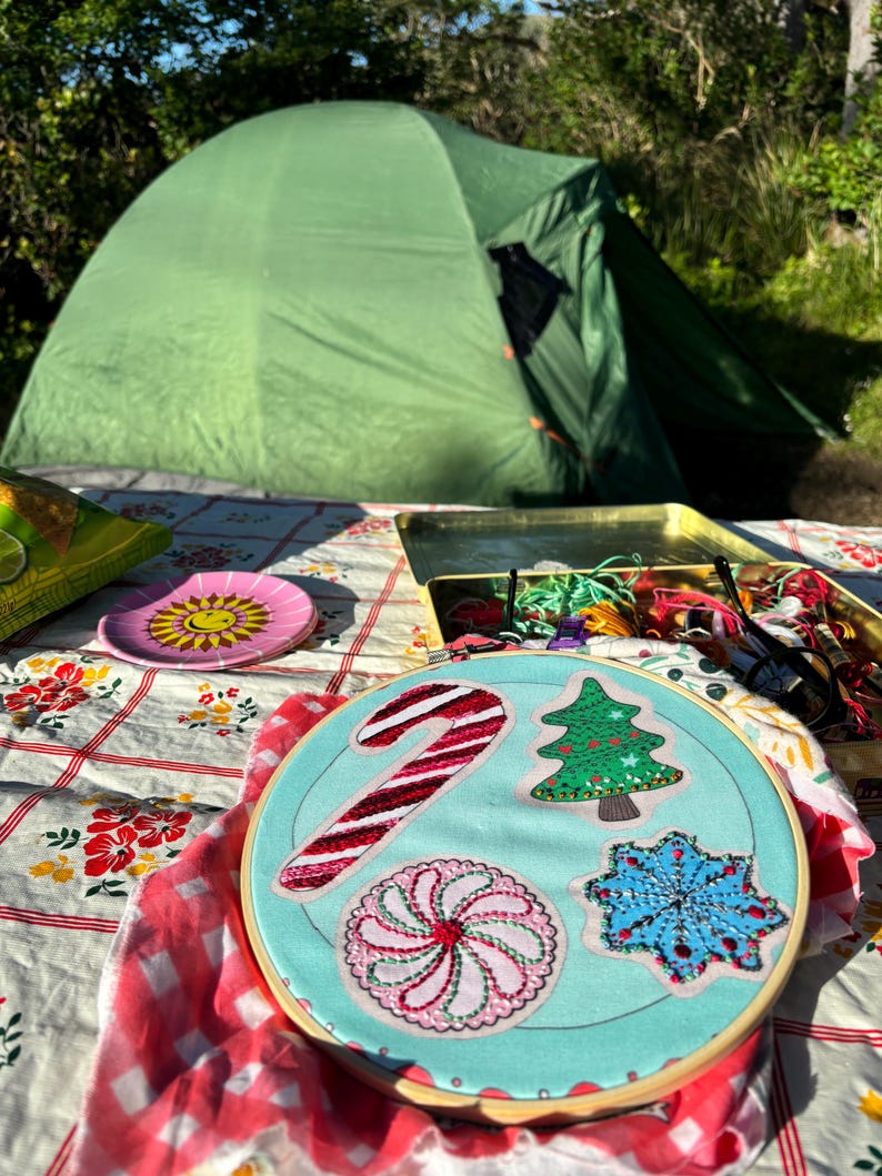 May include: A close-up of a blue embroidery hoop with a candy cane, a Christmas tree, a peppermint swirl, and a snowflake design. The hoop is on a red and white checkered tablecloth.