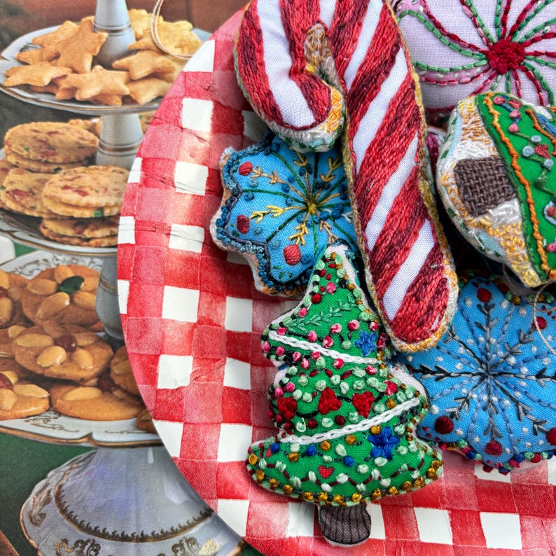 May include: A collection of handmade felt Christmas ornaments on a red and white checkered background. The ornaments include a candy cane, a snowflake, a Christmas tree, and a gingerbread house.