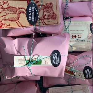 May include: Pink paper packages tied with twine and labeled with "Dropcloth Samplers" and "D.M.C."  The packages are arranged on a green surface.