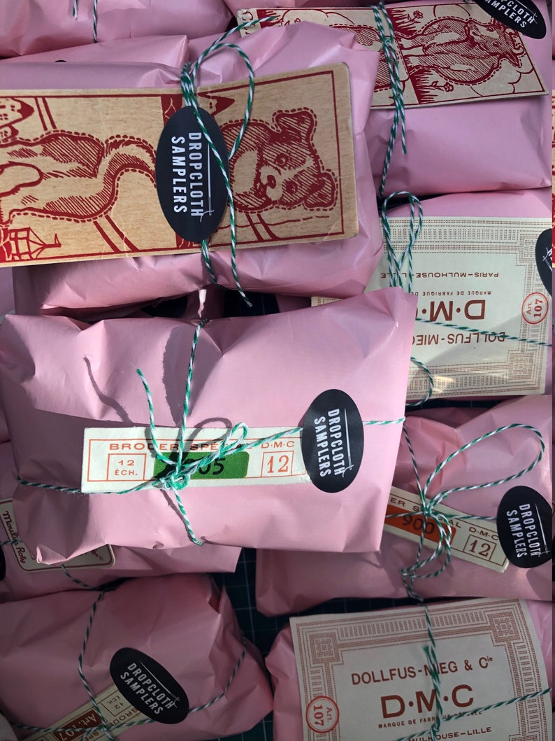 May include: Pink paper packages tied with twine and labeled with "Dropcloth Samplers" and "D.M.C."  The packages are arranged on a green surface.