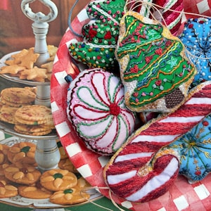 May include: A collection of handmade Christmas ornaments, including a candy cane, a pink and green flower, a green Christmas tree, and a blue snowflake. The ornaments are displayed on a red and white checkered surface.