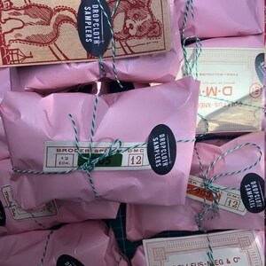 May include: Pink paper packages tied with green and white string. Each package has a label with the text "Dropcloth Samplers" and "D.M.C."  The packages are stacked on a green surface.