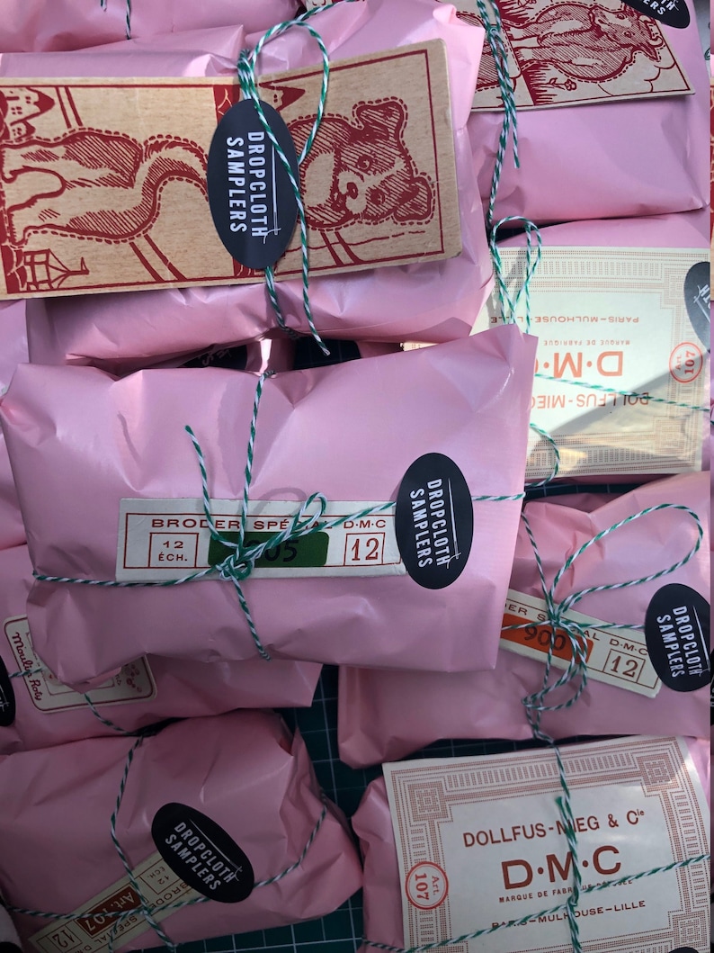 May include: Pink paper packages tied with green and white string. Each package has a label with the text "Dropcloth Samplers" and "D.M.C."  The packages are stacked on a green surface.