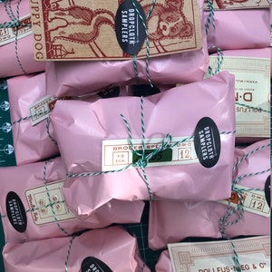 May include: A close-up of several pink packages wrapped with twine and labeled with "Dropcloth Samplers" and "D.M.C." The packages are arranged on a green grid surface.